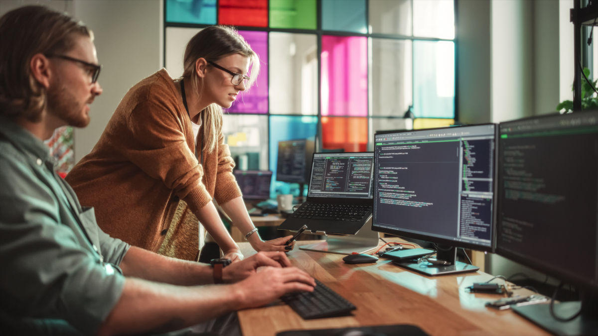 Male data scientist and female developer working on bugs in code on a desktop computer in a creative office